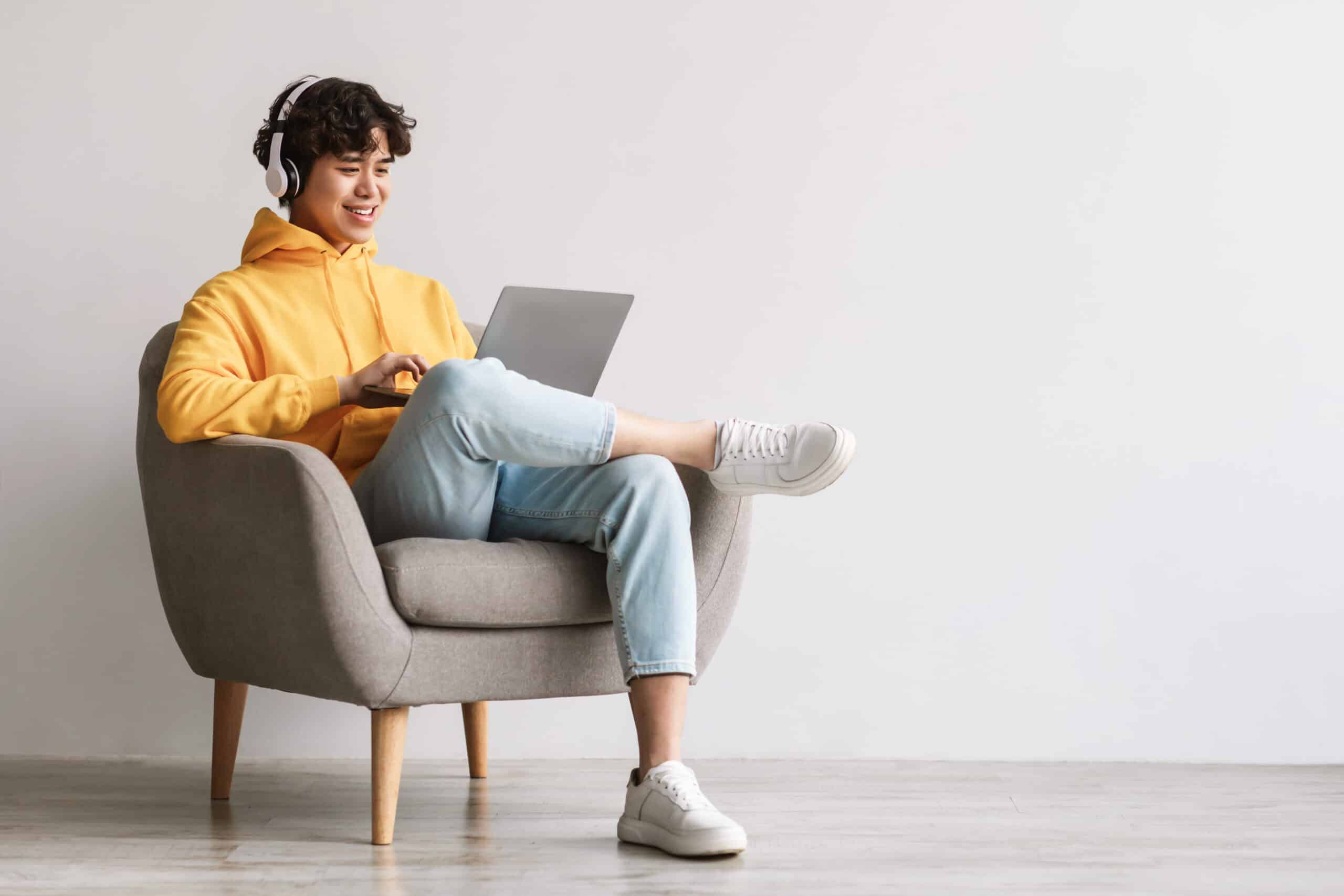 Happy young Asian man using laptop, wearing headphones while sitting in armchair against white wall